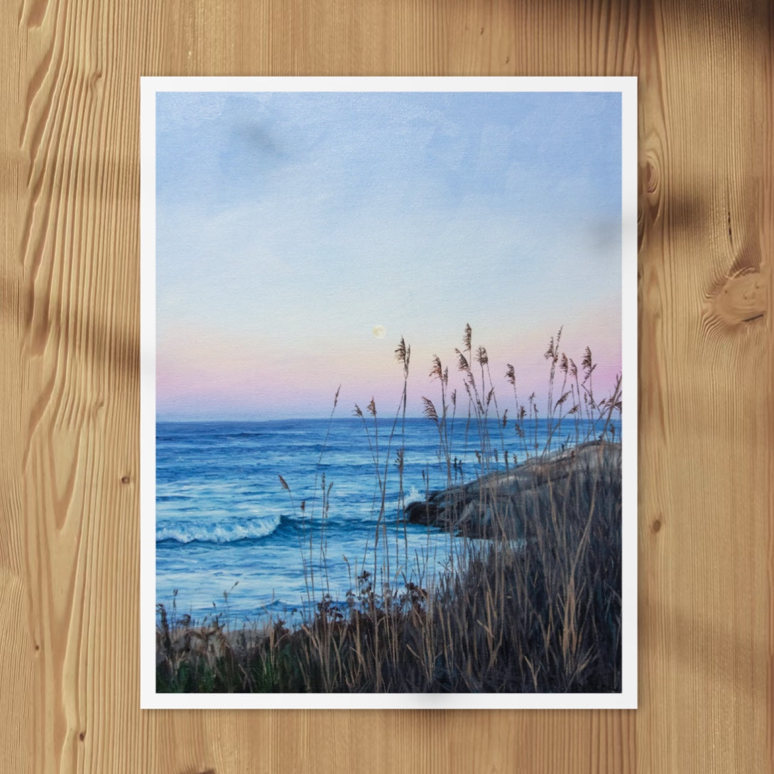 A vertical art print of a coastal seascape at dusk, featuring blue ocean waves, a pale pink and blue sky, and beach grasses in the foreground, displayed on a light wood background.