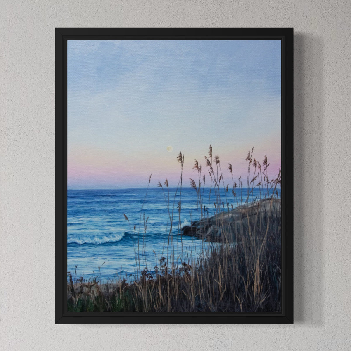 A framed landscape art print depicting a coastal scene at dusk, with blue ocean waves, a pale sky with soft pink and lavender tones, and tall dry grasses in the foreground, mounted on a light gray textured wall.