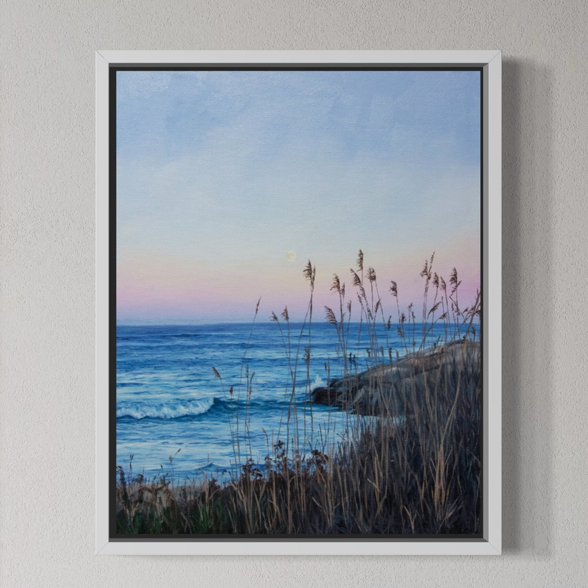 A framed coastal landscape painting hanging on a textured light gray wall. The artwork shows blue ocean waves, a pale pink and blue sky, and dark grasses in the foreground.