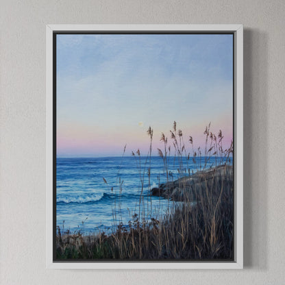 A framed coastal landscape painting hanging on a textured light gray wall. The artwork shows blue ocean waves, a pale pink and blue sky, and dark grasses in the foreground.