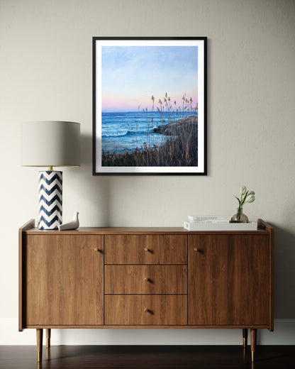 A framed vertical landscape art print depicting a coastal beach at dusk, styled on a light gray wall above a mid-century modern wooden sideboard with a lamp and books.