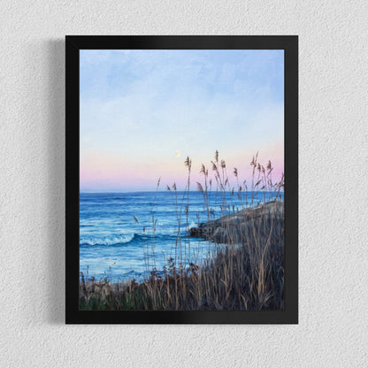 A framed coastal landscape print hangs on a textured white wall. The artwork shows blue ocean waves, a pale blue and pink sky, and dark grasses in the foreground.