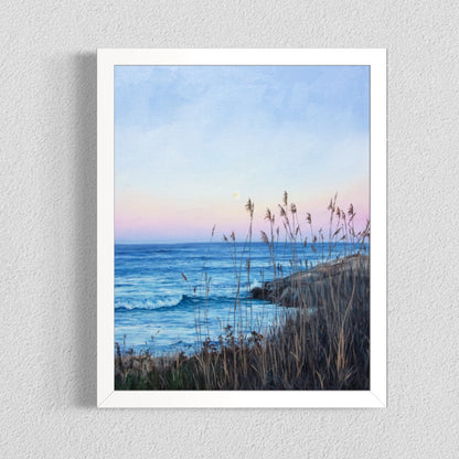 A framed art print depicting a serene coastal landscape at dusk, featuring blue ocean waves, a pale pink and blue sky, and dry grasses in the foreground, mounted on a textured light gray wall.