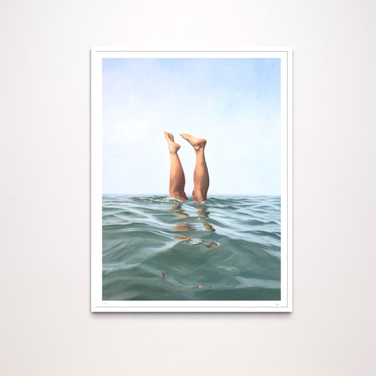 A framed art print featuring a surreal photograph of a person's legs extending upward from the ocean water under a pale blue sky, displayed on a light gray wall.