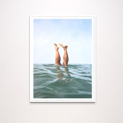 A framed art print featuring a surreal photograph of a person's legs extending upward from the ocean water under a pale blue sky, displayed on a light gray wall.