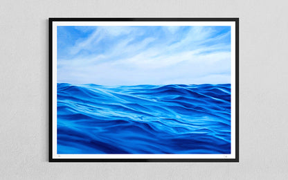 A framed art print depicting a vibrant blue ocean under a cloudy sky, mounted on a light gray textured wall.