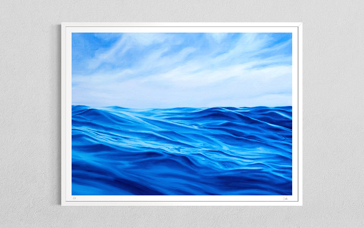 A framed art print depicting a vibrant blue ocean under a cloudy sky, mounted on a light gray textured wall.