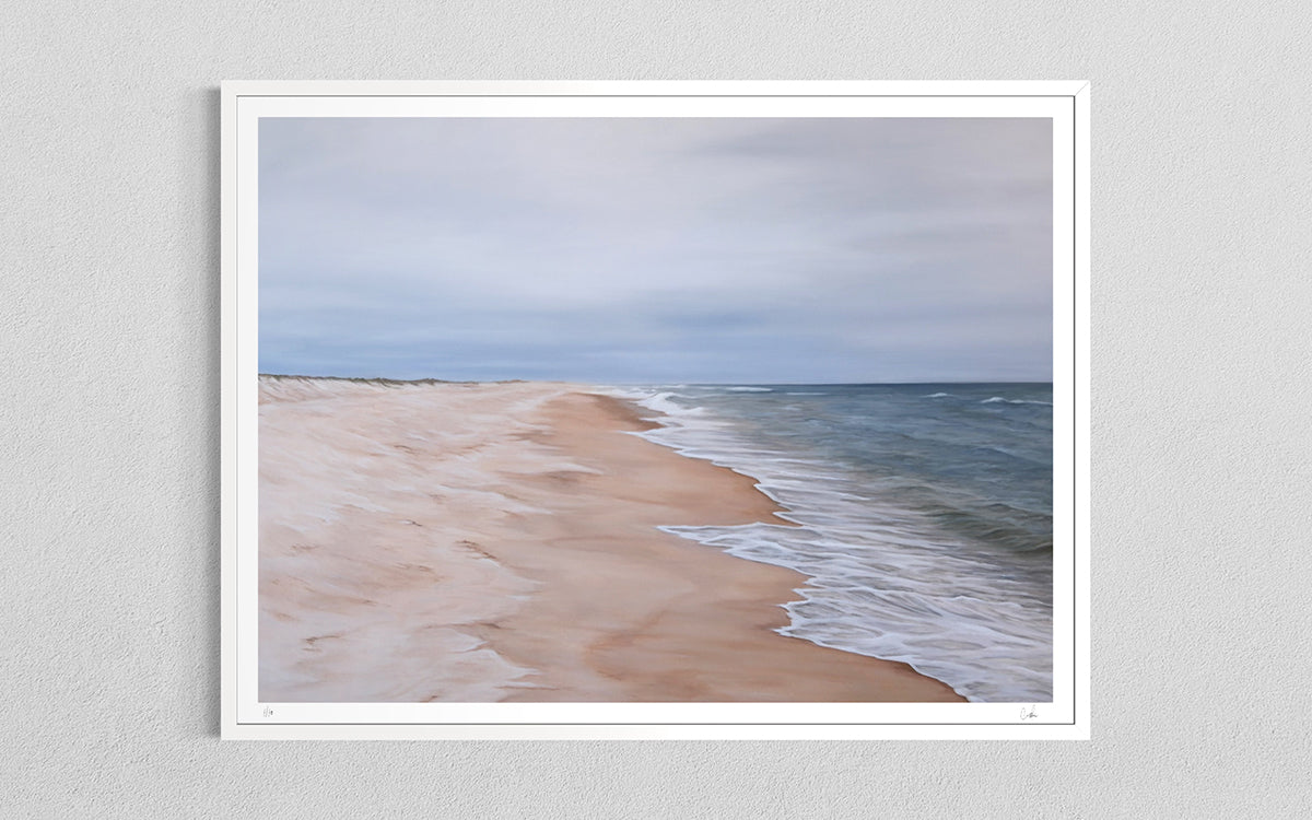 A framed art print depicting a serene beach scene with soft sand, gentle waves, and a cloudy sky, mounted on a light gray textured wall.