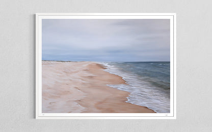 A framed art print depicting a serene beach scene with soft sand, gentle waves, and a cloudy sky, mounted on a light gray textured wall.