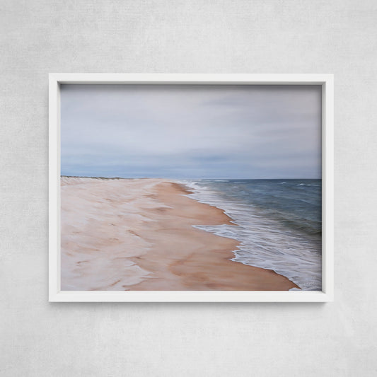 A framed fine art paper print of a coastal scene with a sandy beach and calm sea, displayed on a wall.