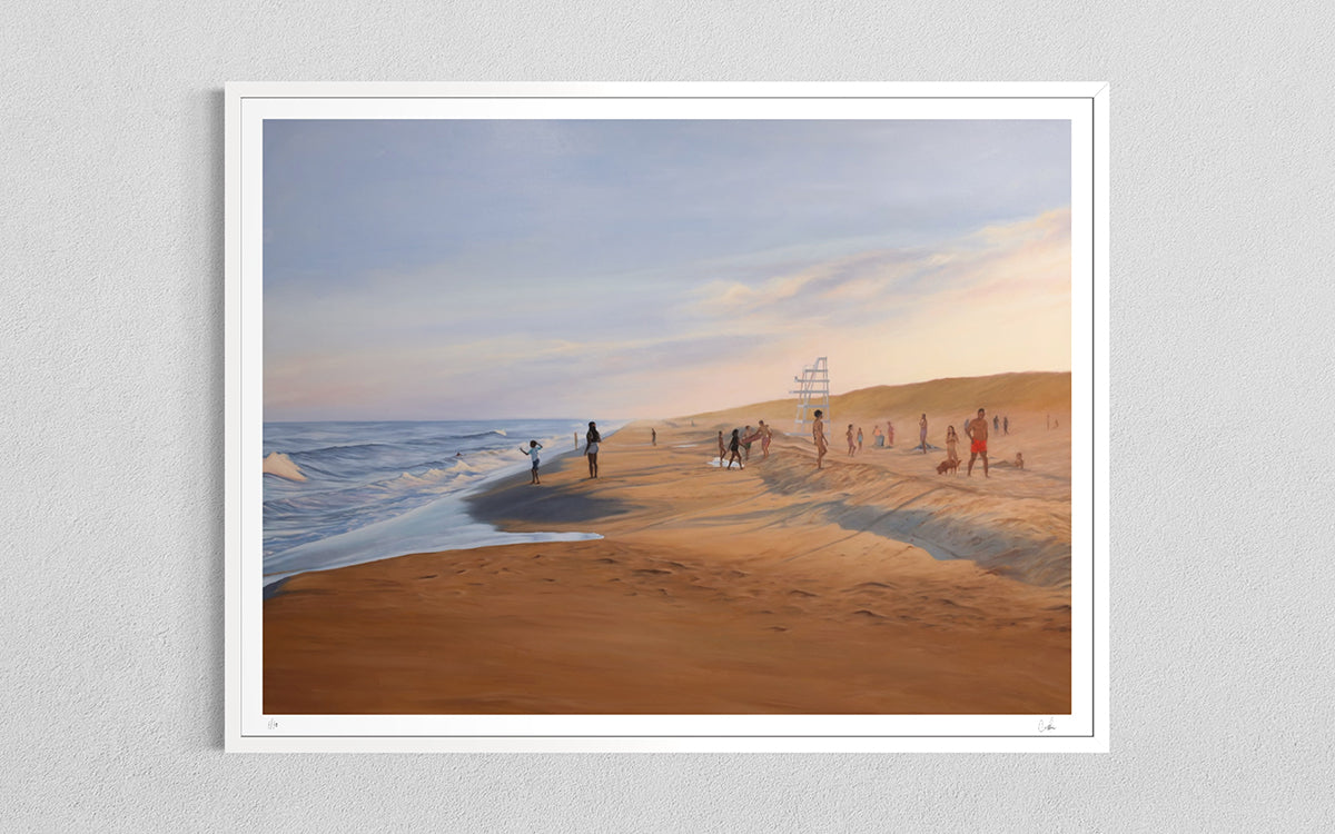 A framed art print depicting a beach scene with sand dunes, ocean waves, and small figures of people, mounted on a light gray textured wall.