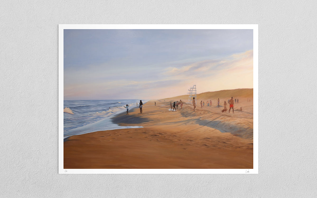 A framed art print depicting a beach scene with sand dunes, ocean waves, and small figures of people, displayed against a light gray textured wall.